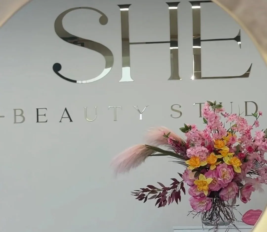 SHE beauty studio siena ar zelta logo un rozā ziedu kompozīciju, skatīta apaļajā spogulī